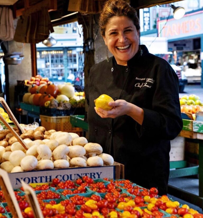 A smiling woman in a black chef's coat stands by a vibrant market stall. She holds a lemon and is surrounded by fresh produce, including tomatoes and mushrooms. The setting is lively, with people and market signs in the background.