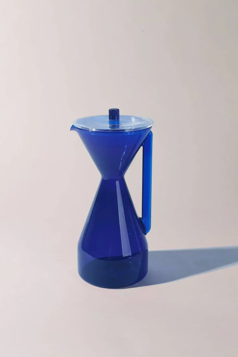 A cobalt blue, hourglass-shaped pitcher with a lid and handle is placed against a plain, light background, casting a soft shadow to the side.