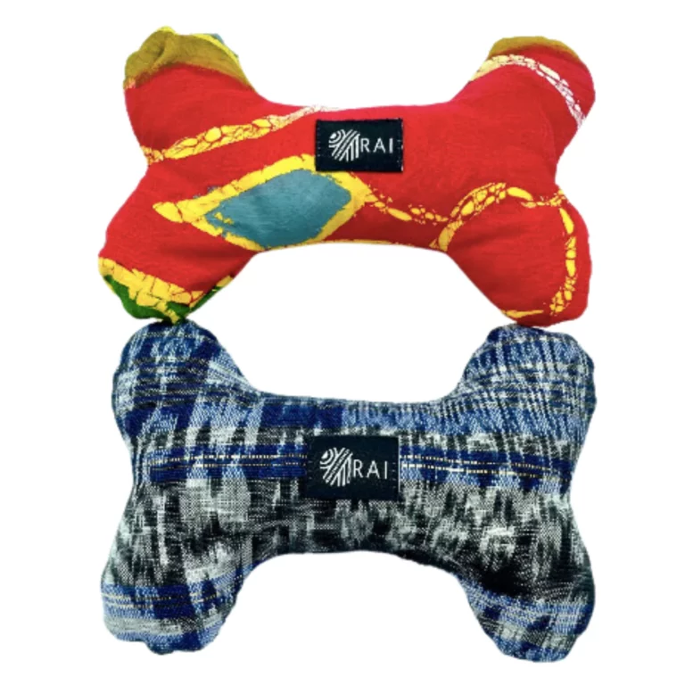 Two dog bone-shaped toys are shown. The top toy is red with colorful patterns, while the bottom toy is blue with plaid and abstract designs. Both feature a small branded label in the center.