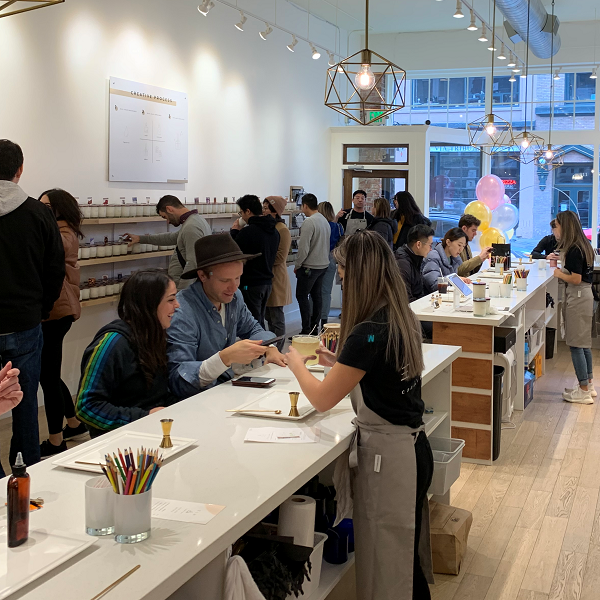 People in a candle-making workshop, seated at long white tables, crafting candles. A person helps another with wax pouring. The room is bright with modern decor, and various candle supplies are visible. Balloons and a window are in the background.