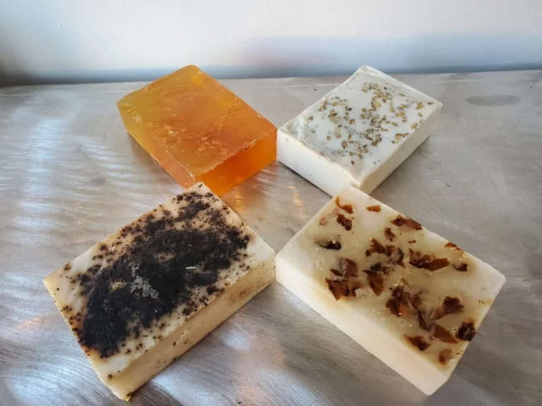 Four handmade soap bars are arranged on a metal surface. Each has a distinct appearance, with various natural ingredients visible: one is translucent amber, another has dark speckles, and two contain small light and dark pieces.