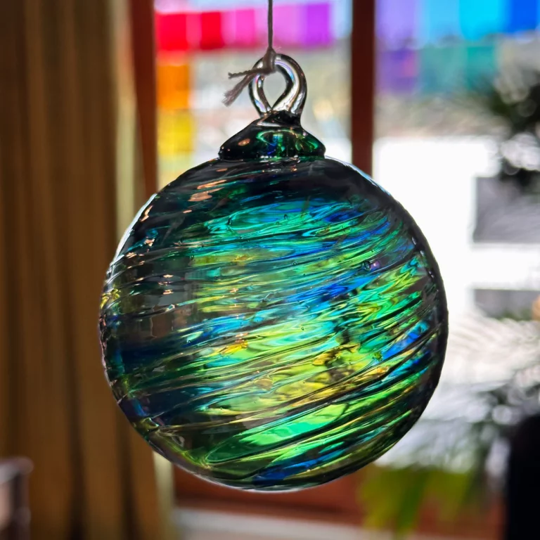 A colorful, translucent glass ornament hangs in front of a window. The ornament swirls with shades of green, blue, and hints of yellow. The window behind displays a blurred mosaic of bright, multicolored squares.