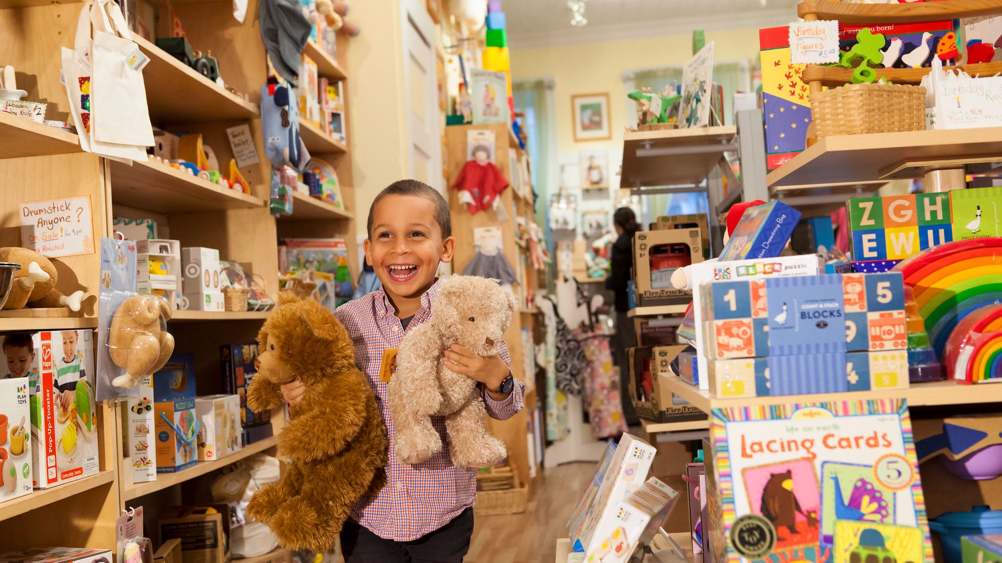 A child excitedly holds 2 stuffed bears in a toy store, surrounded by other toys for sale.